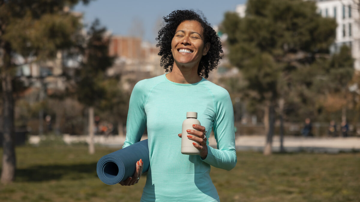 Mulher sorridente vestindo blusa esportiva azul, segurando um tapete de yoga enrolado e uma garrafa d’água em um parque, com árvores e prédios ao fundo.