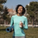 Mulher sorridente vestindo blusa esportiva azul, segurando um tapete de yoga enrolado e uma garrafa d’água em um parque, com árvores e prédios ao fundo.