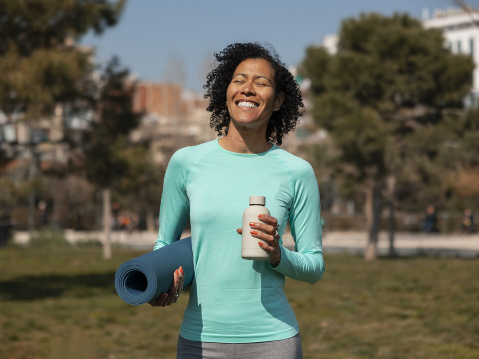 Mulher sorridente vestindo blusa esportiva azul, segurando um tapete de yoga enrolado e uma garrafa d’água em um parque, com árvores e prédios ao fundo.