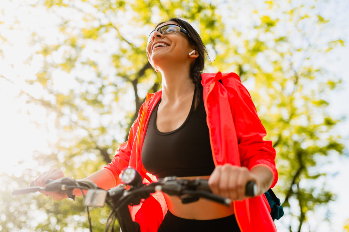 Mulher sorridente usando óculos escuros e jaqueta esportiva vermelha, andando de bicicleta em um parque com árvores ao fundo. Ilustrar um estilo de vida ativo e saudável ao ar livre