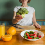 Mulher apresentando um brócolis como símbolo de alimentação saudável. A mesa está repleta de opções nutritivas, como suco de laranja, laranjas frescas, salada de espinafre e tomate, além de uma garrafa de azeite.