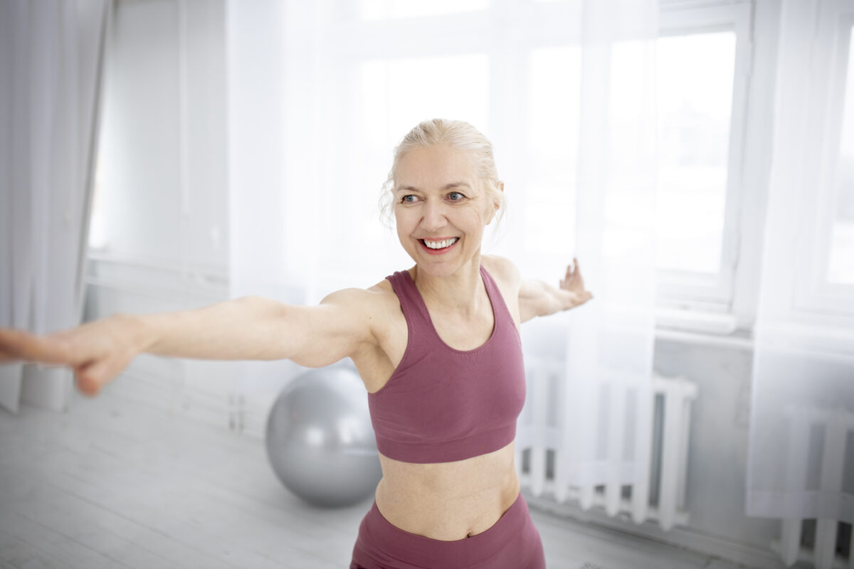 Mulher sorridente com roupa esportiva rosa realizando uma pose de yoga com os braços abertos, dentro de uma sala bem iluminada.