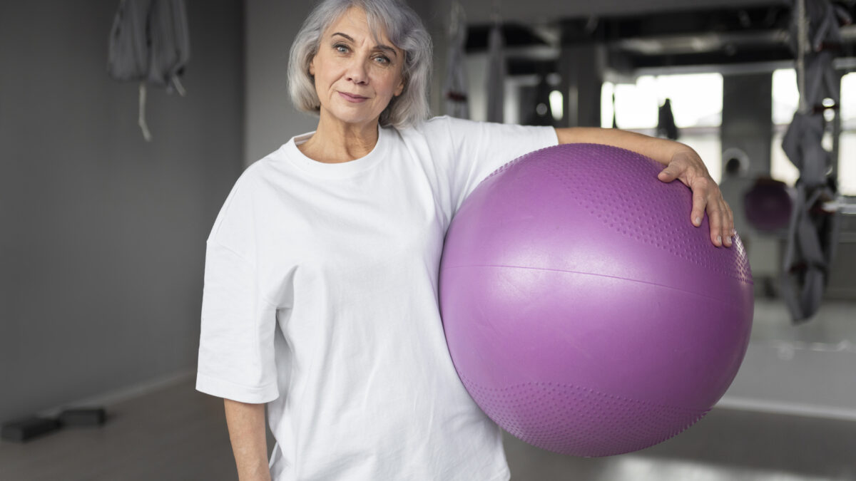 Mulher grisalha, de meia-idade, segurando uma bola de pilates em uma academia.