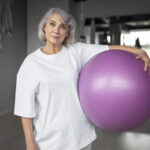 Mulher grisalha, de meia-idade, segurando uma bola de pilates em uma academia.