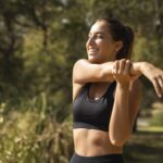 Mulher jovem sorridente, vestindo roupa de treino preta, se alonga ao ar livre em um ambiente verde e ensolarado.